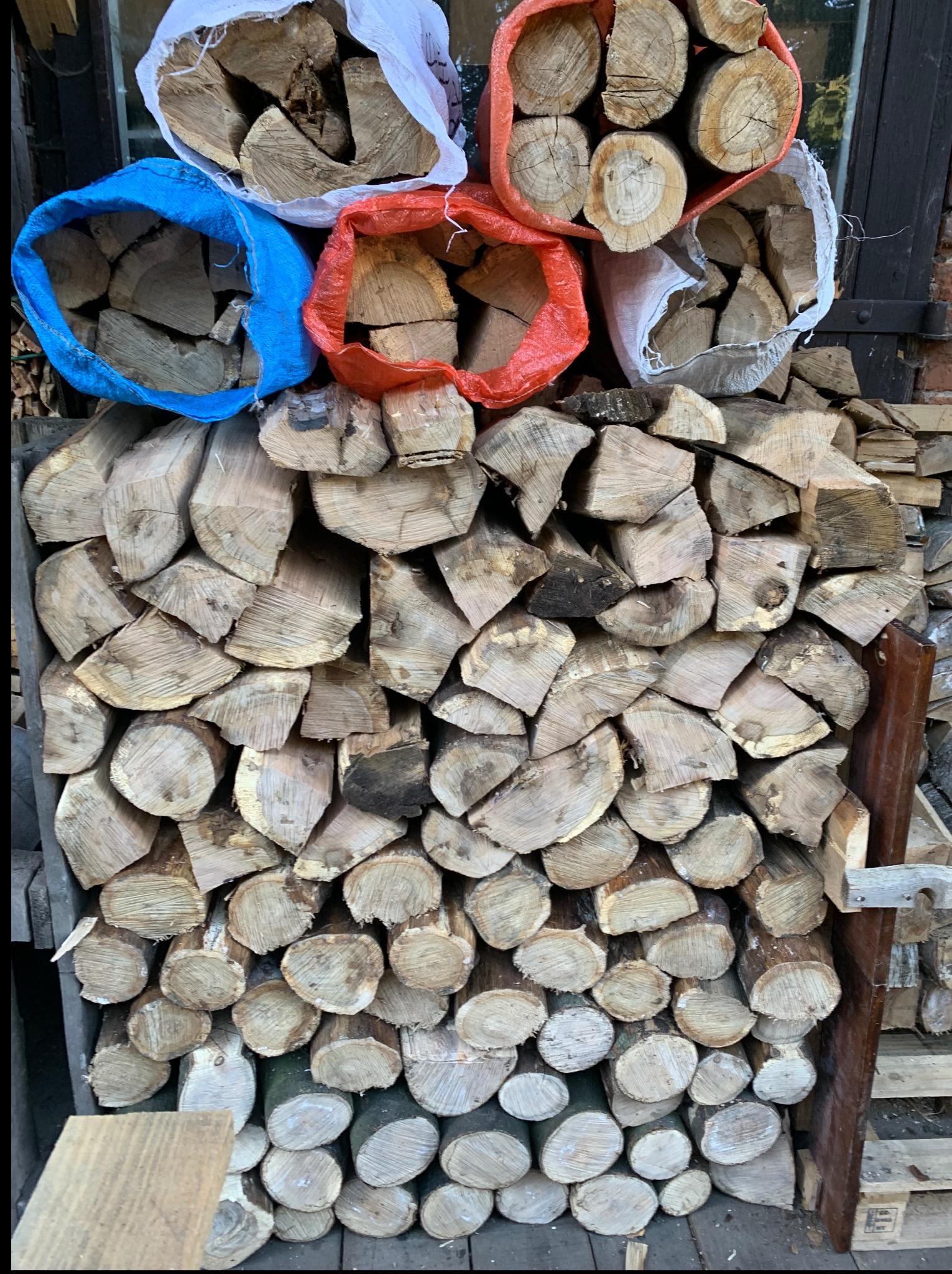 Ułożone drewno kominkowe, część w workach (niebieski, czerwony, biały), część luzem. Widoczne słoje drewna i kora. Selekcja drewna do kominka.