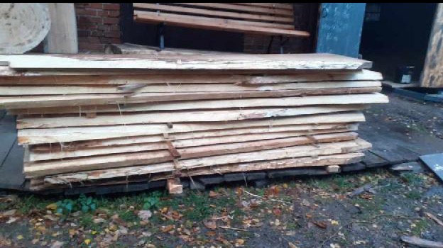 Stos tarcicy z jasnego drewna, zabezpieczony metalowymi taśmami, leżący na drewnianym podłożu na zewnątrz. W tle ławka i ceglany budynek.