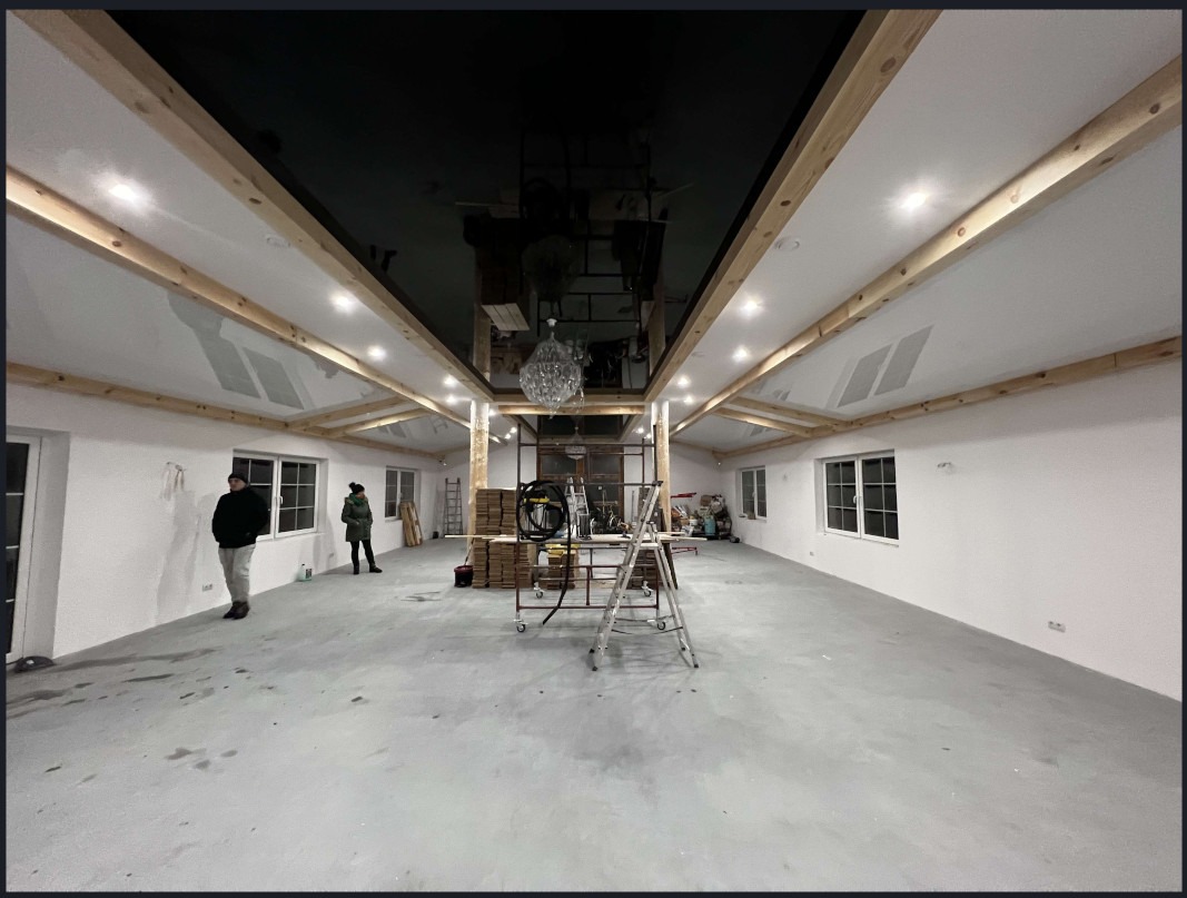 Duże wnętrze z błyszczącym, czarnym sufitem napinanym, drewnianymi belkami i oświetleniem punktowym. W tle drabina i materiały budowlane, sugerujące etap wykończenia.