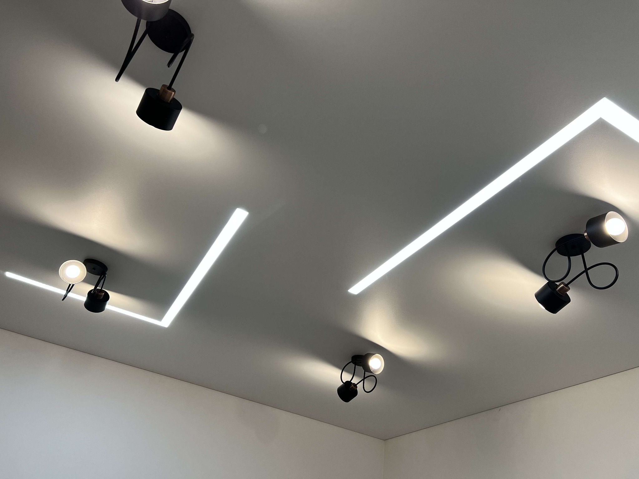 Biały sufit z nowoczesnym oświetleniem: liniowe LED i czarne reflektory punktowe. Minimalistyczny design, ciepłe światło. Ujęcie z dołu.