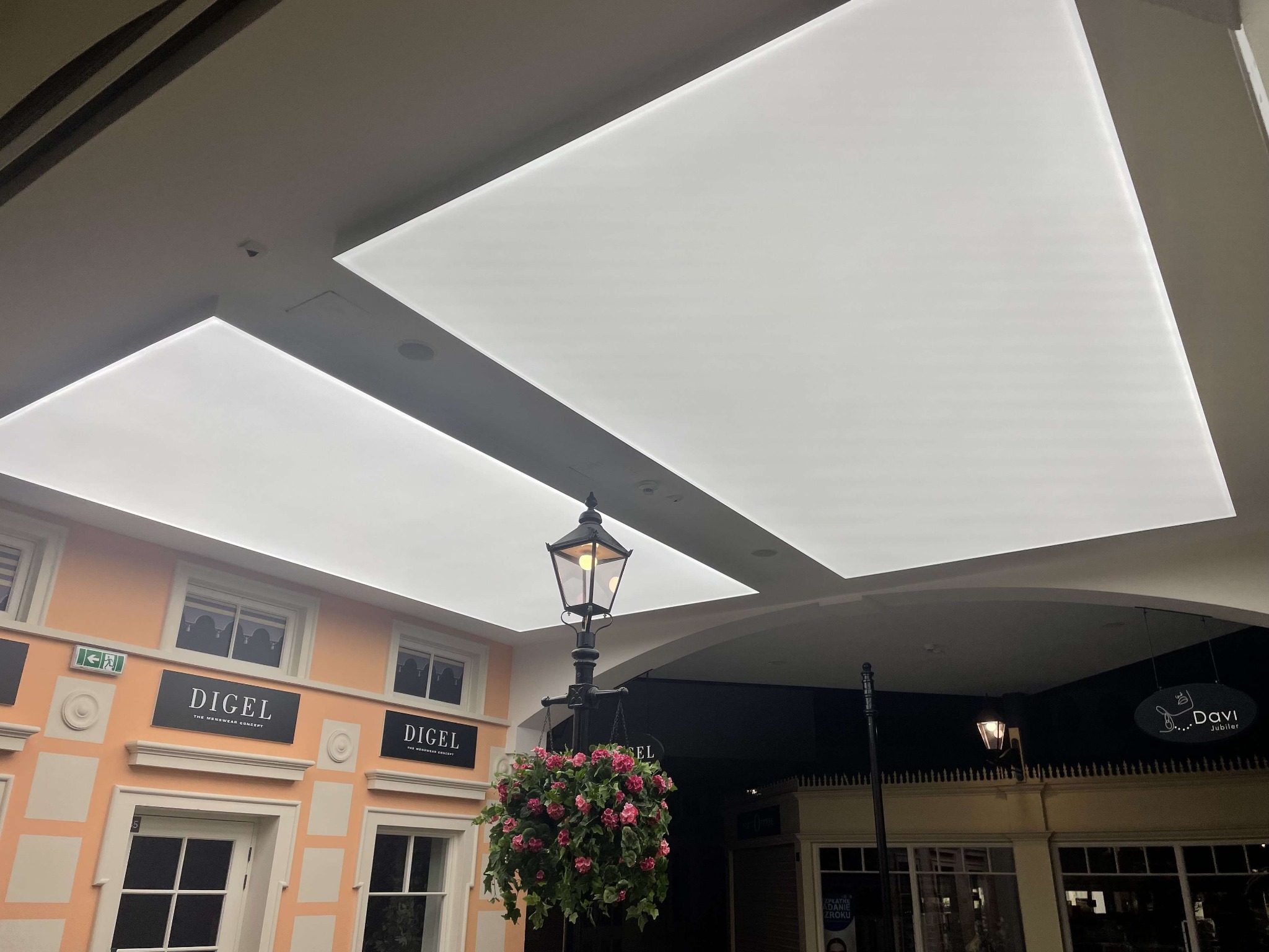 Białe panele sufitowe podświetlone w galerii handlowej, z lampą uliczną i kwiatami. Widoczne fasady sklepów Digel i Davi Jubiler.