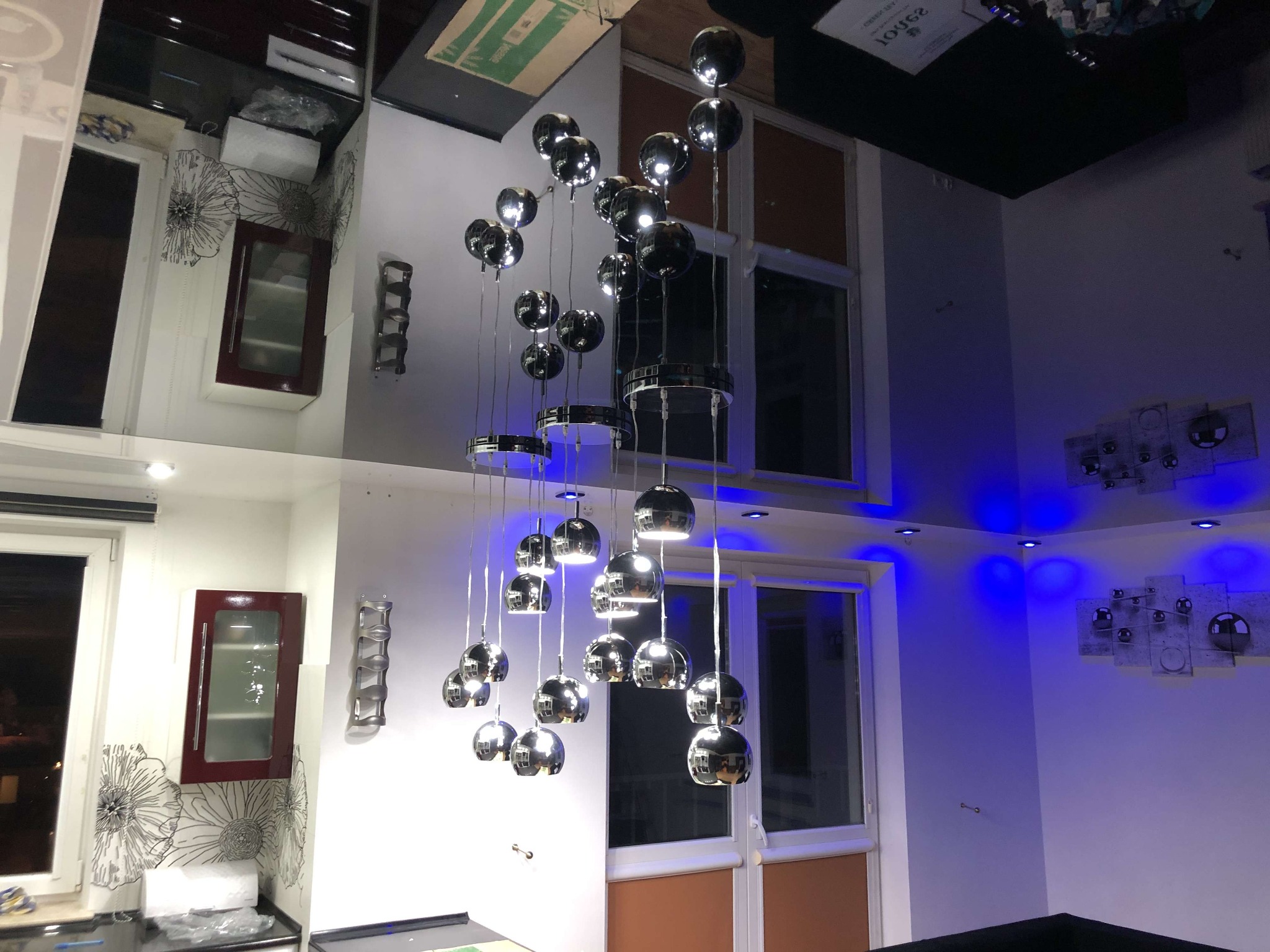 Nowoczesny salon z lustrzanym sufitem, oświetleniem LED i wiszącą lampą z chromowanymi kulami. Widoczna zabudowa kuchenna w tle i dekoracje ścienne.