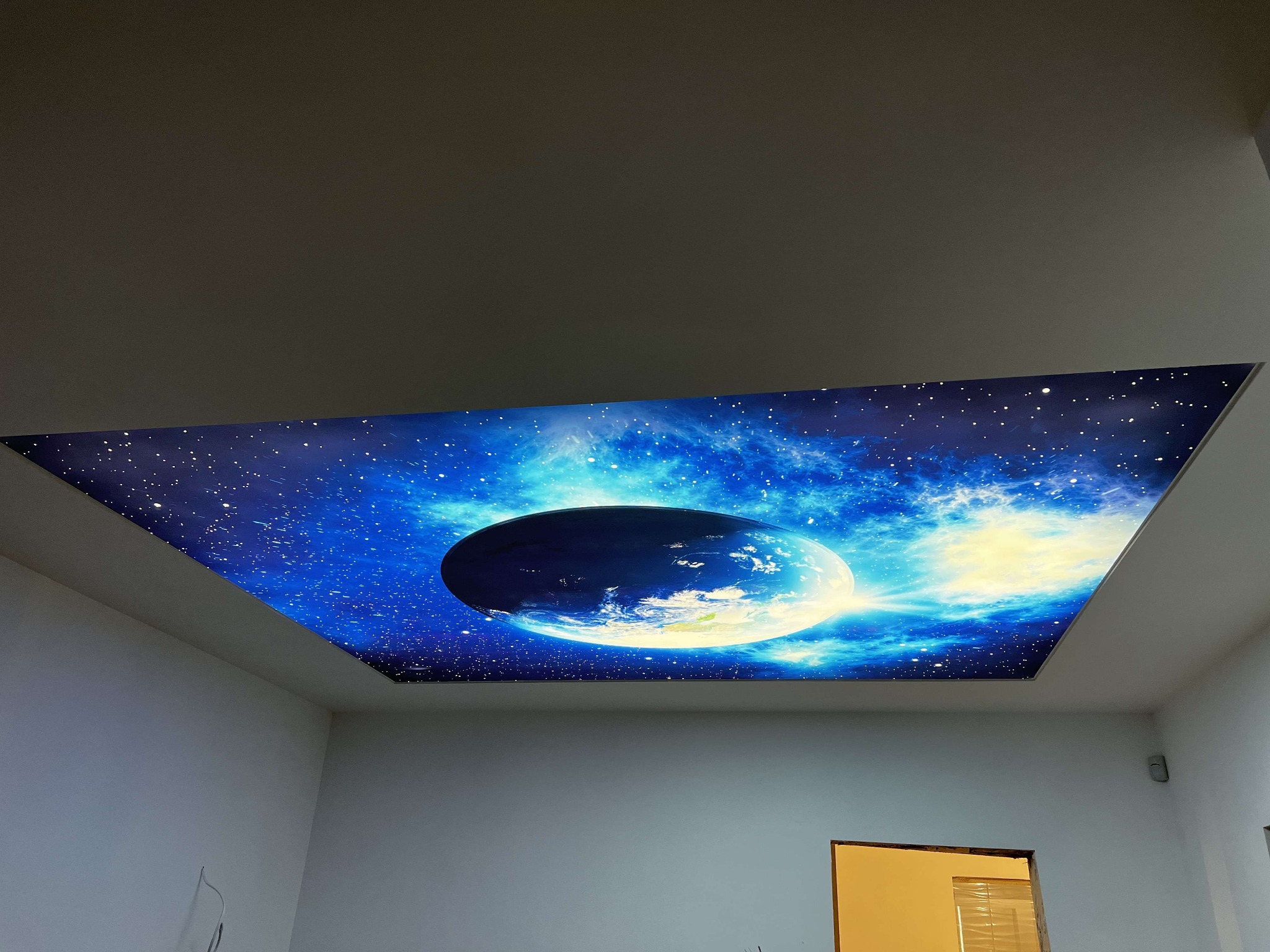 Nowoczesny sufit napinany z motywem kosmosu i planety Ziemi, oświetlony od wewnątrz, w białym pomieszczeniu. Efektowne wykończenie wnętrza.