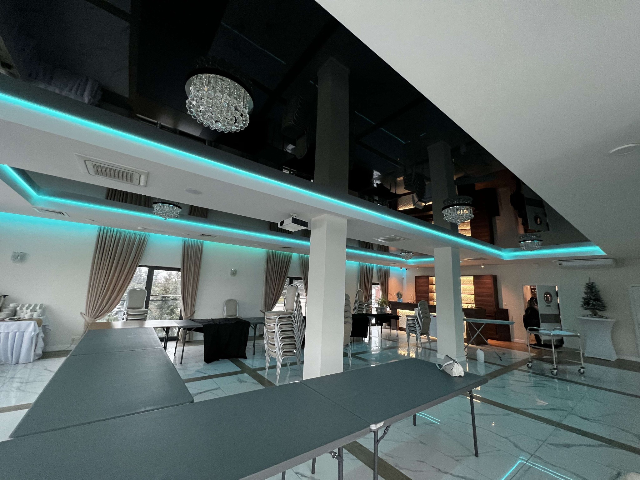 Sala bankietowa z czarnym, błyszczącym sufitem napinanym, oświetleniem LED i kryształowymi żyrandolami, przygotowana do uroczystości. Widoczne stoły i krzesła.