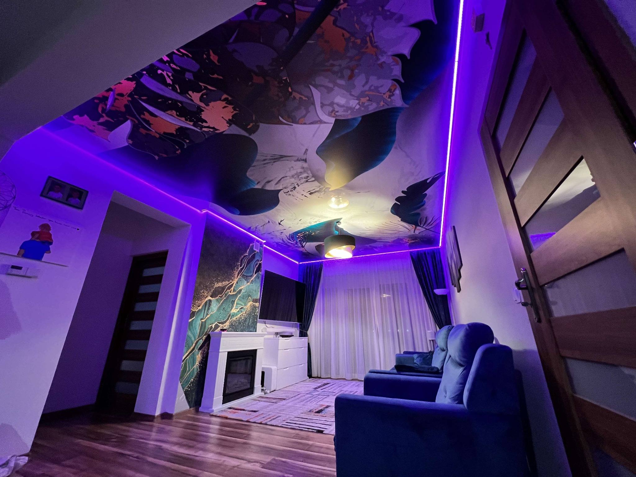 Nowoczesny salon z sufitem napinanym z abstrakcyjnym wzorem, oświetlonym fioletowym LED. Wnętrze dopełnia kominek, TV i niebieska sofa.