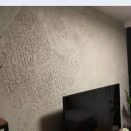 Dekoracyjna ściana w salonie z szarą fakturą i ornamentami geometrycznymi. Na pierwszym planie telewizor i roślina doniczkowa.