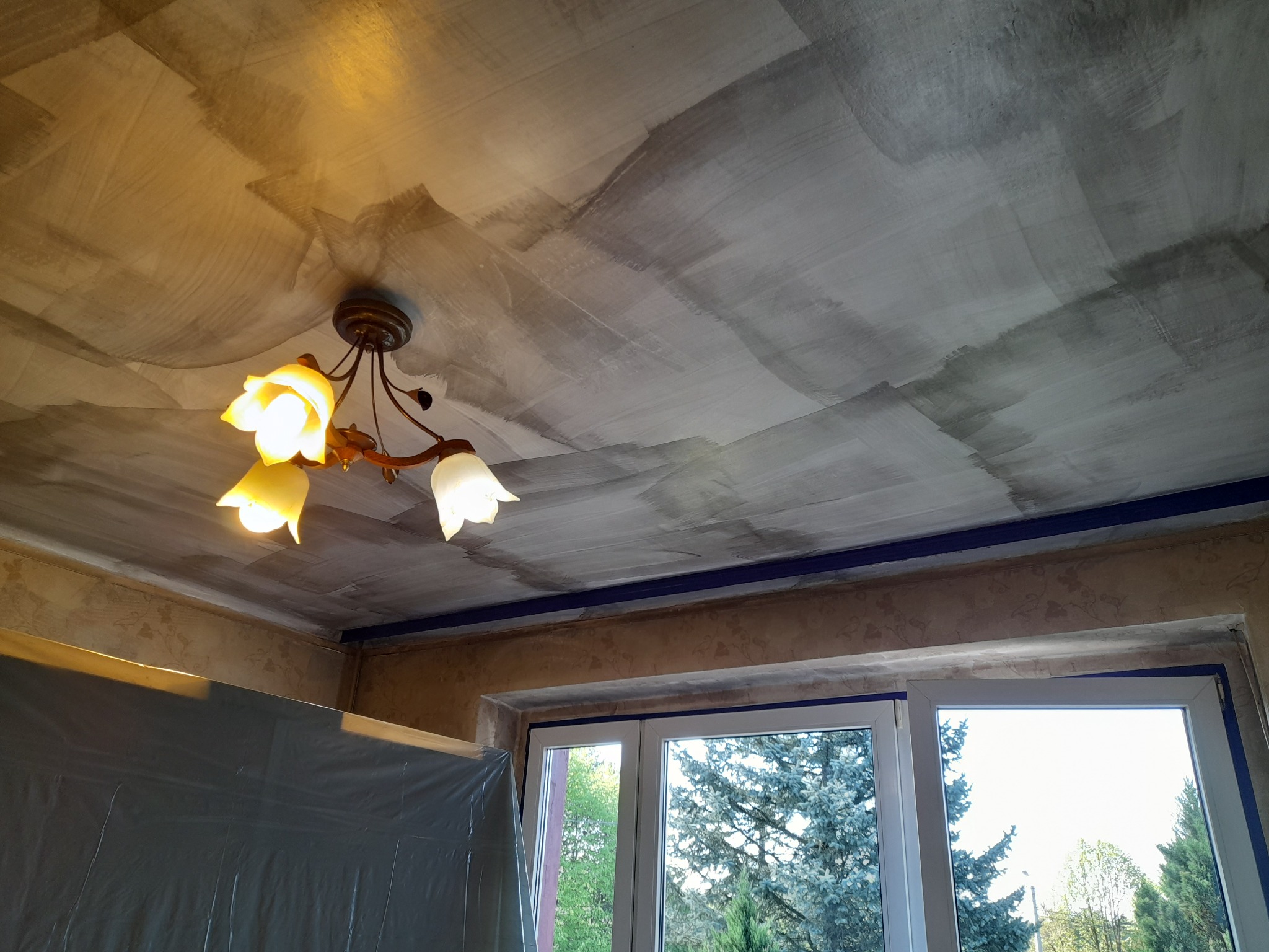 Malowany sufit w szarych tonach, z widoczną fakturą i lampą w stylu retro. Zabezpieczone taśmą malarską okno z widokiem na zieleń.