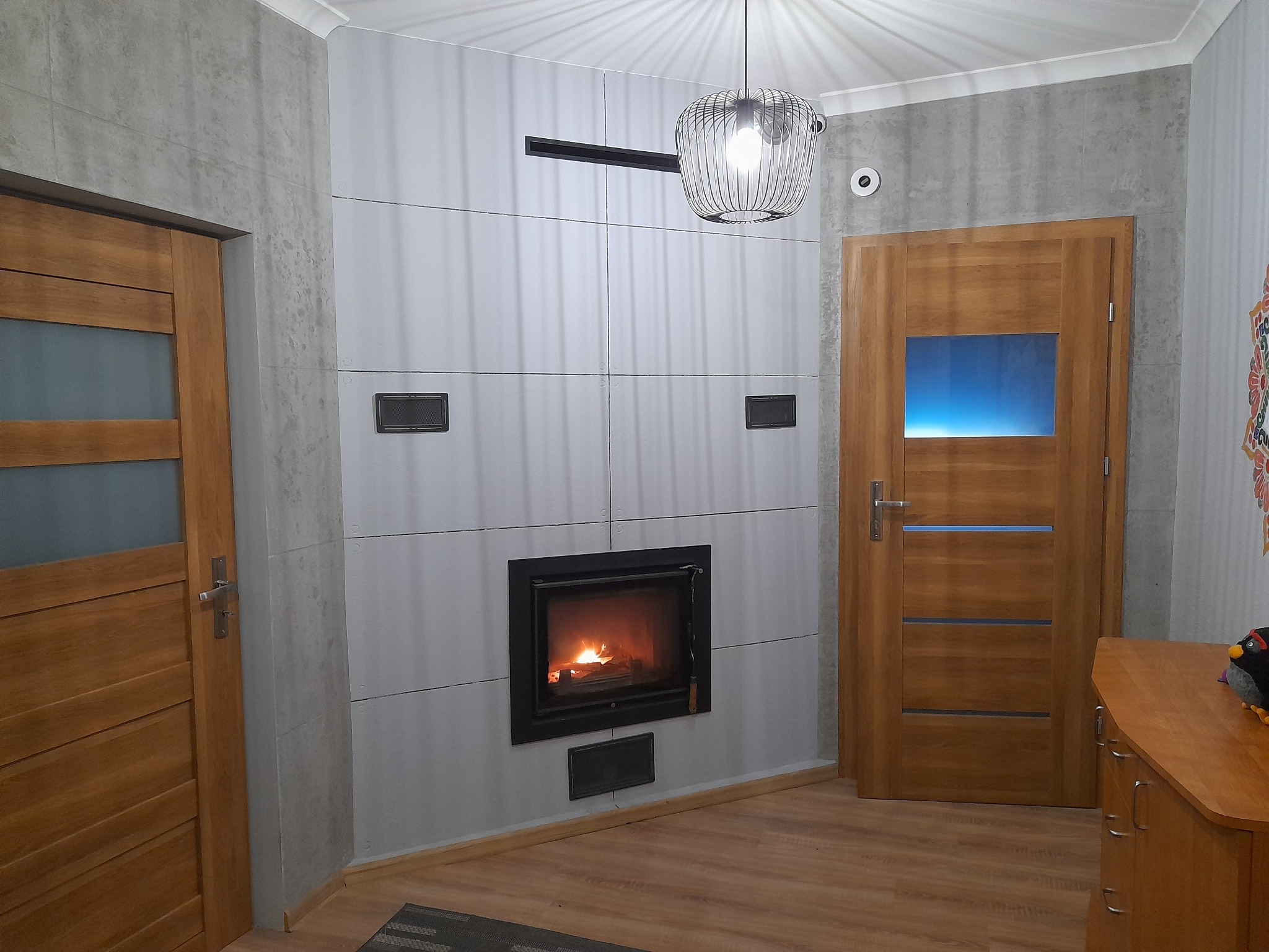 Nowoczesny salon z kominkiem obudowanym płytami imitującymi beton, drewnianymi drzwiami i podłogą. Wisząca lampa z metalowym ażurem.