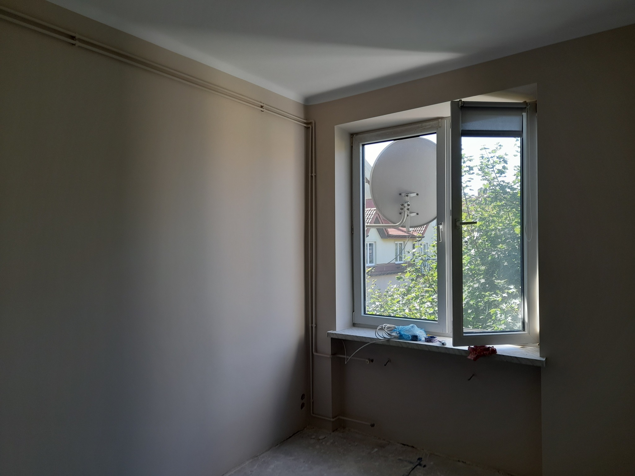 Jasny pokój po malowaniu ścian na beżowo, z otwartym oknem i widoczną anteną satelitarną za oknem. Rury grzewcze wzdłuż ściany. Świeżo pomalowane ściany.