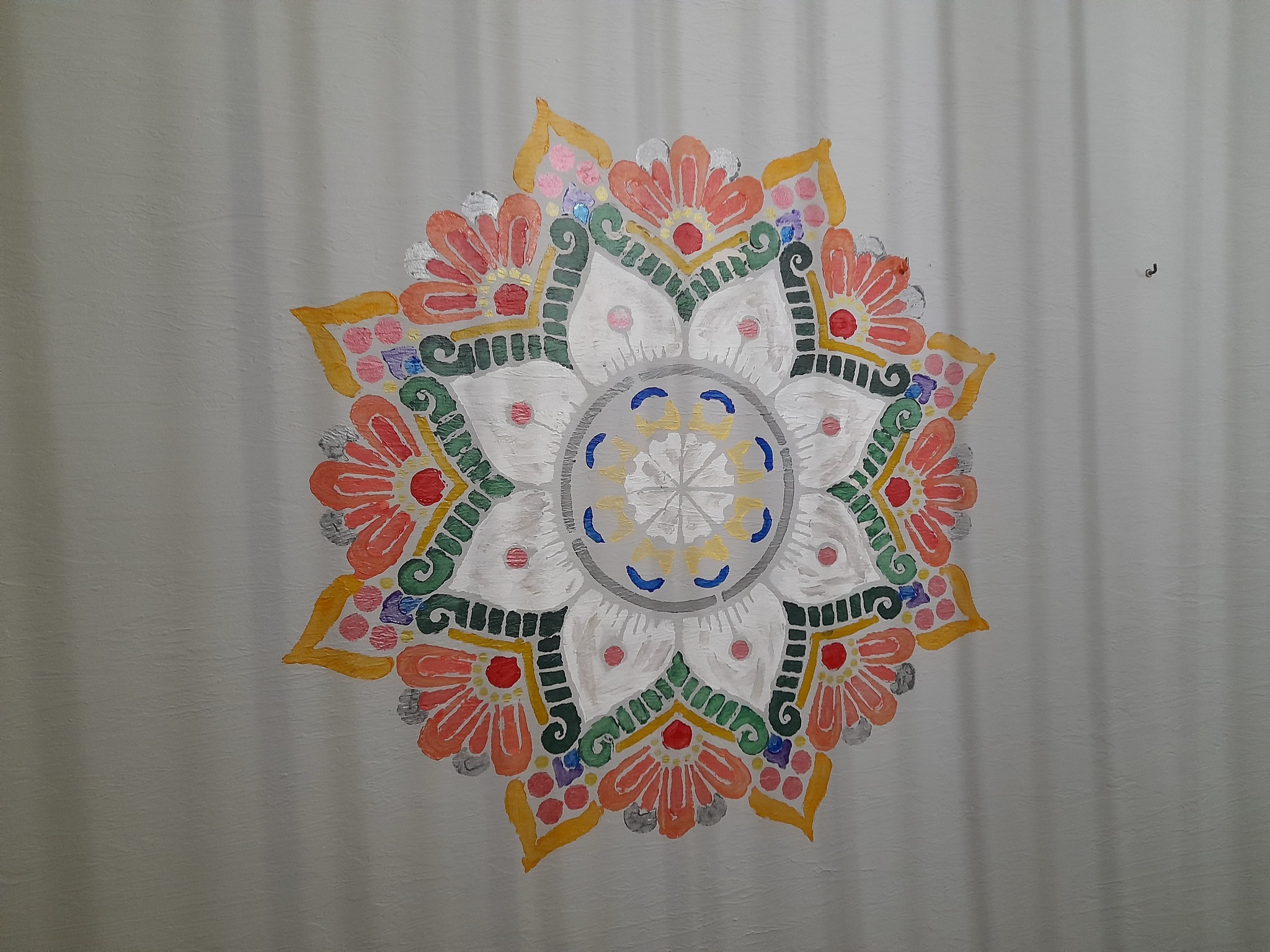Ręcznie malowana mandala w odcieniach bieli, pomarańczy, żółci i zieleni na jasnoszarej ścianie z widoczną fakturą. Dekoracja ścienna.
