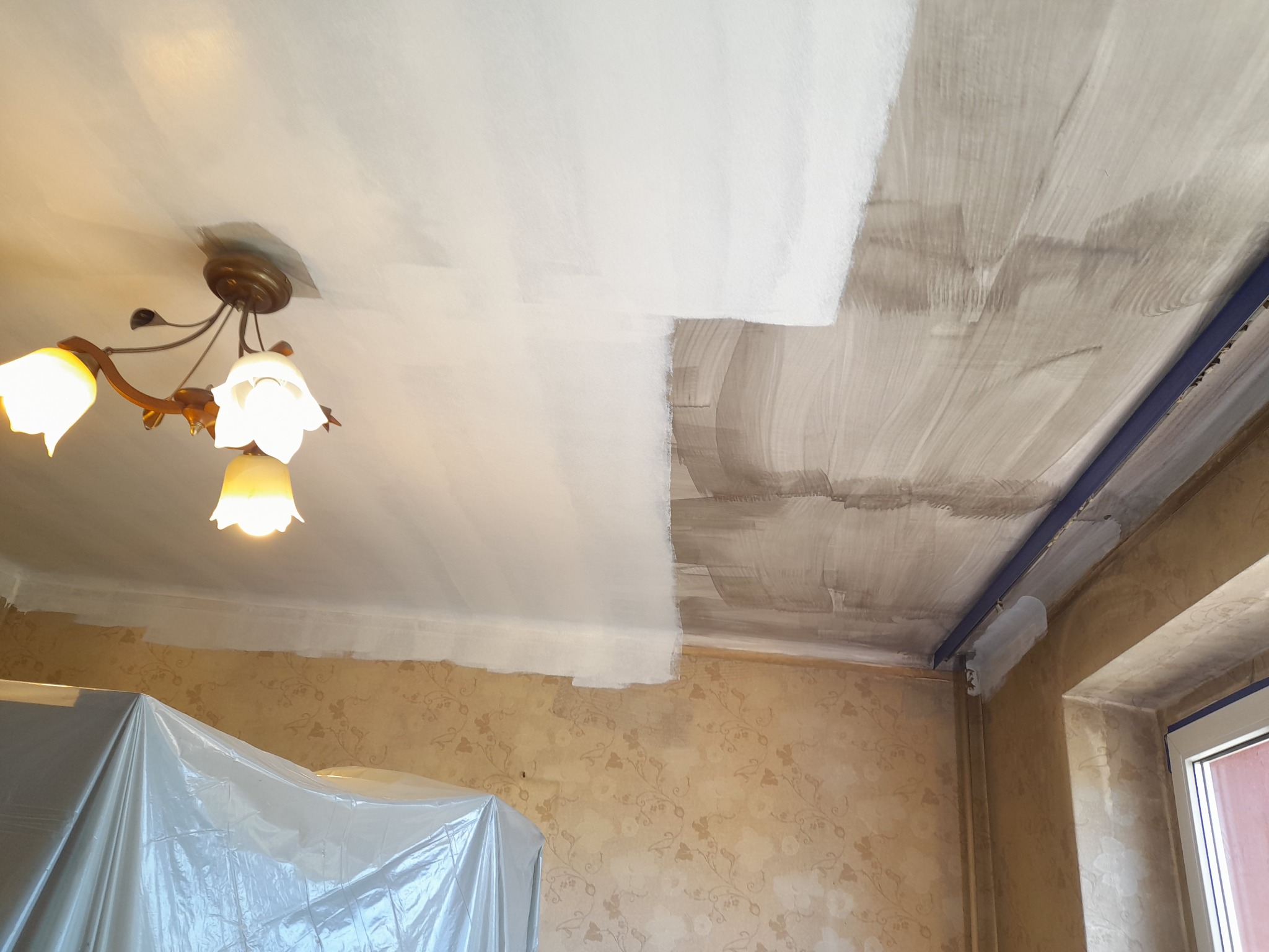 Częściowo pomalowany sufit z widoczną lampą i zabezpieczoną folią ścianą, ukazujący postęp prac. Fragment sufitu z widocznymi pociągnięciami pędzla.