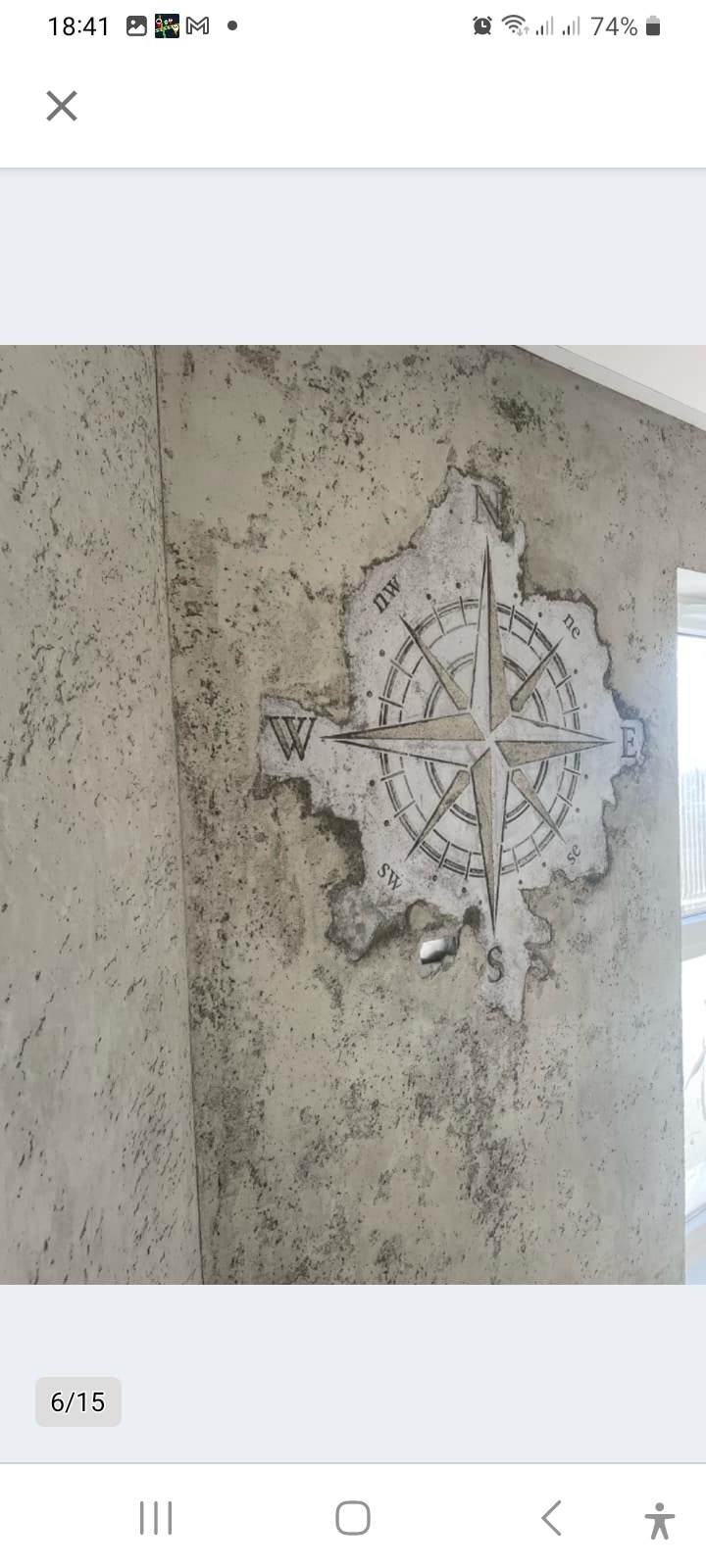 Dekoracyjna ściana z efektem starego tynku i wkomponowaną w niego grafiką kompasu z zaznaczonymi kierunkami świata. Stylizacja rustykalna, ściana w odcieniach szarości.