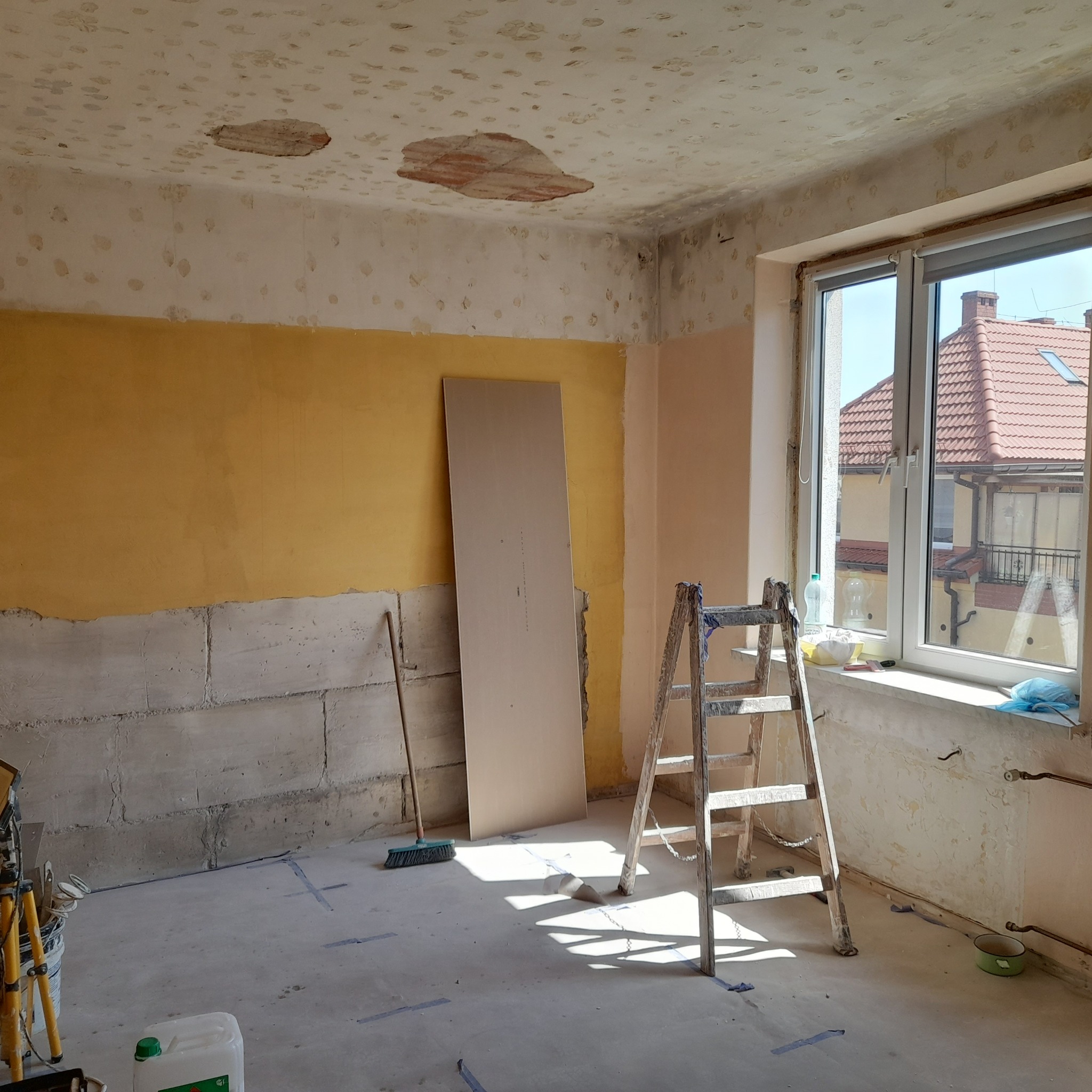 Pokój w trakcie prac: żółta ściana, częściowo usunięta stara powłoka, drabina, płyta gipsowa oparta o ścianę. Widoczne uszkodzenia sufitu i ślady po taśmie malarskiej.