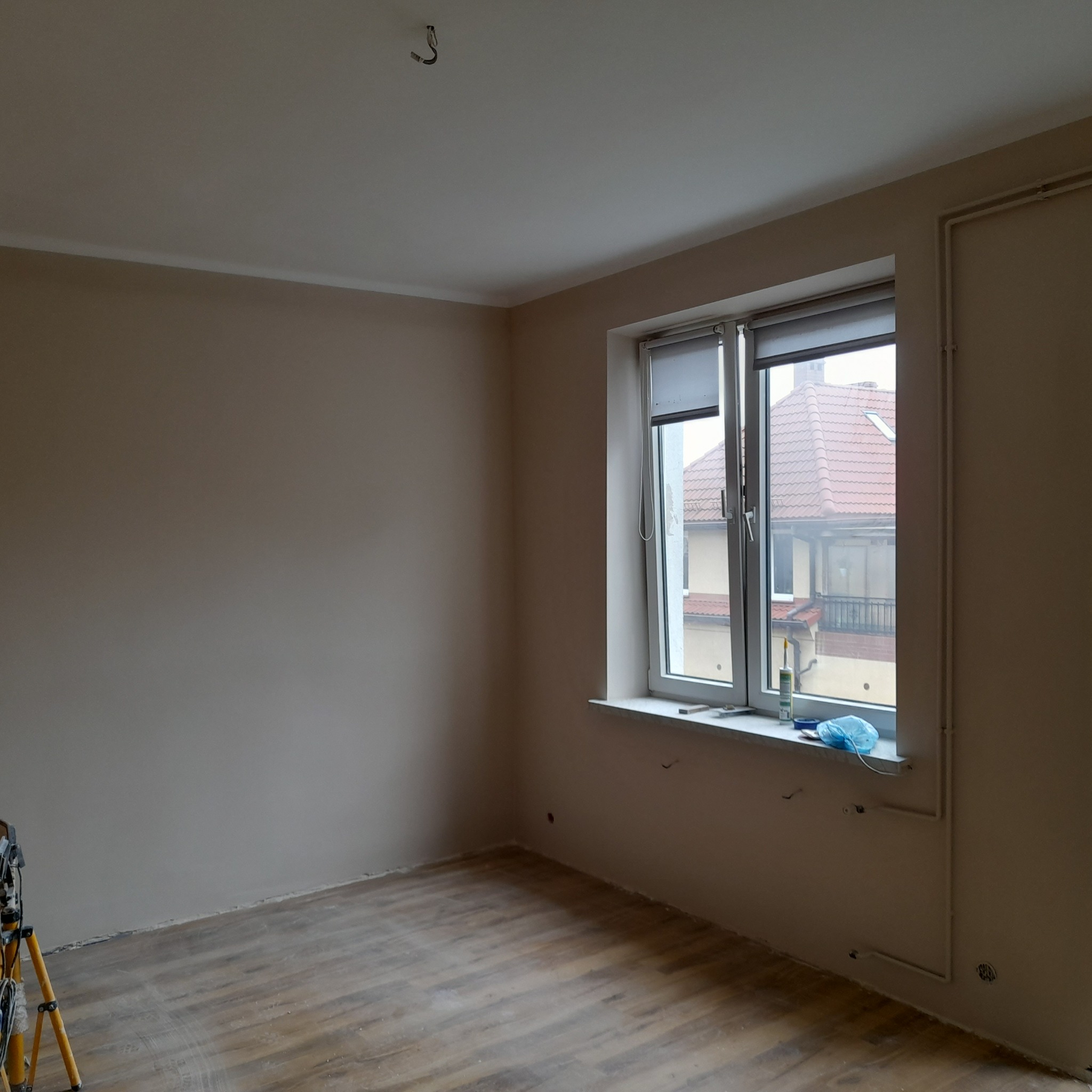 Jasny pokój po remoncie: świeżo pomalowane ściany w odcieniach beżu, drewnopodobna podłoga, białe okno z widokiem na zabudowę. Na parapecie narzędzia i folia ochronna.