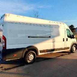 ComfortDrive Sanckler - Biały furgon dostawczy z przesuwnymi drzwiami bocznymi, zaparkowany na asfaltowej drodze w słoneczny dzień. Widok z boku, tło stanowią budynki i drzewa.