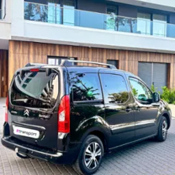 Czarny Citroen Berlingo z naklejką #transport zaparkowany przed nowoczesnym budynkiem z balkonami ze szklanymi balustradami. Widok z tyłu na kostce brukowej.