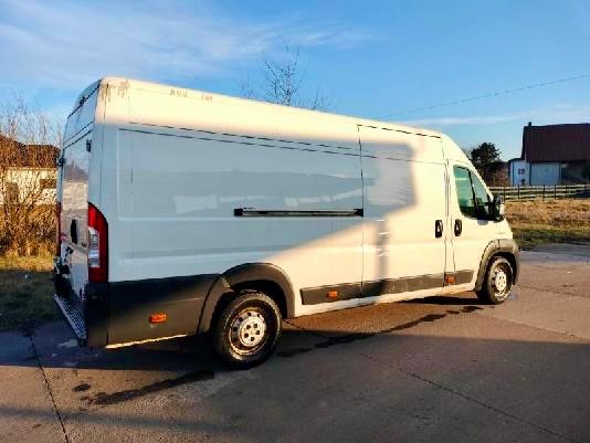 Biały furgon dostawczy z przesuwnymi drzwiami bocznymi, zaparkowany na asfaltowej drodze w słoneczny dzień. Widok z boku, tło stanowią budynki i drzewa.