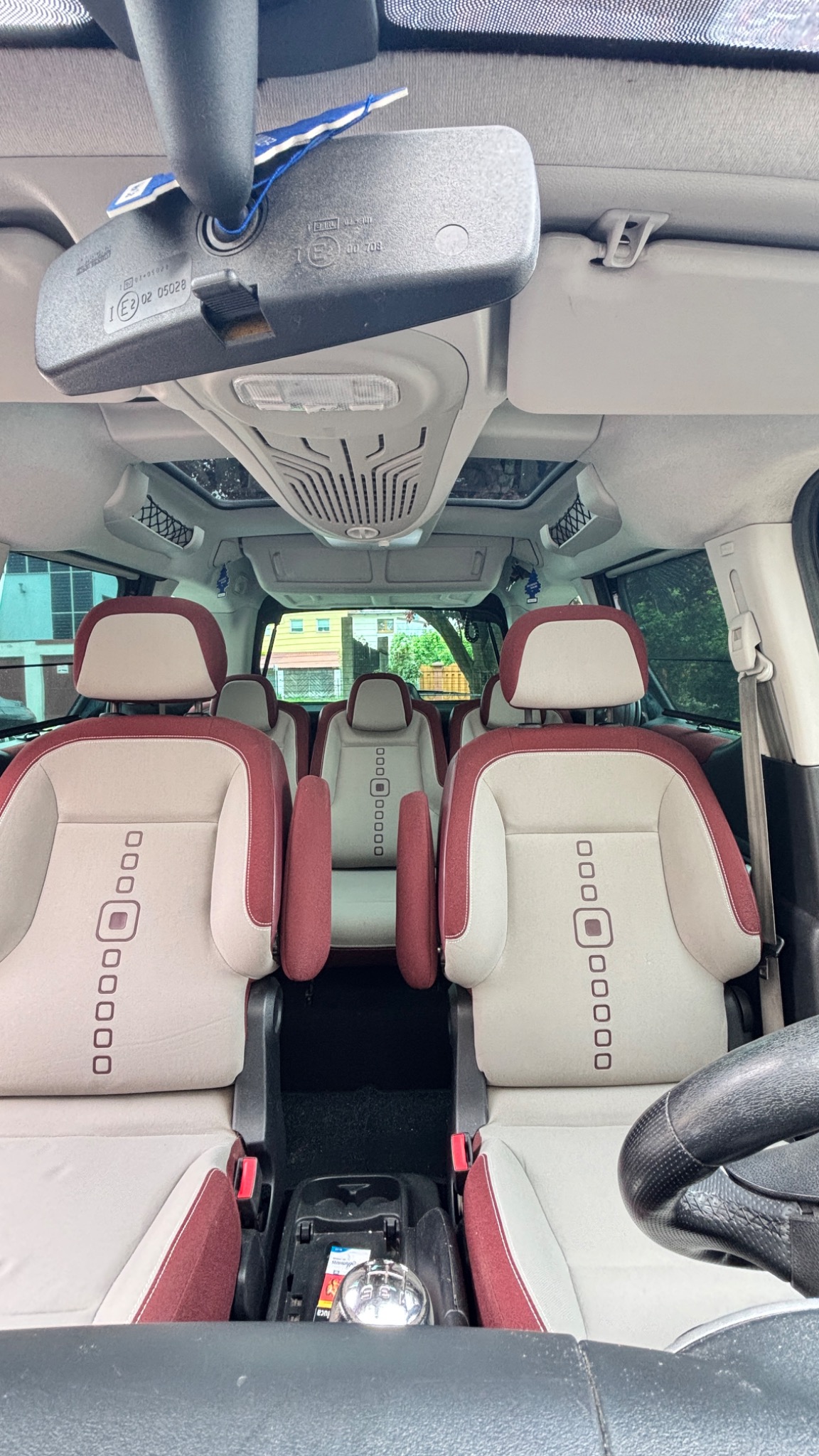 Wnętrze przestronnego, wieloosobowego auta z jasną tapicerką i czerwonymi akcentami. Widoczne fotele, lusterko wsteczne i fragment deski rozdzielczej. Panoramiczny dach.