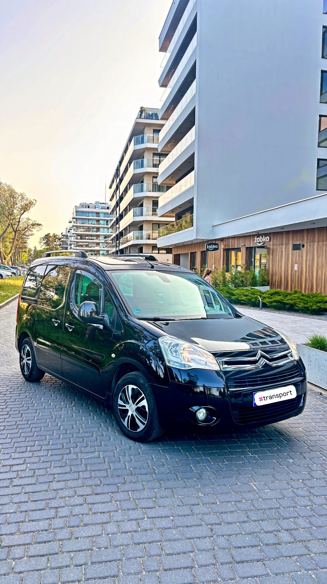 Czarny van Citroen Berlingo na tle nowoczesnego budynku z balkonami i sklepem Żabka. Auto zaparkowane na brukowanej ulicy. Widoczny napis #transport na tablicy.