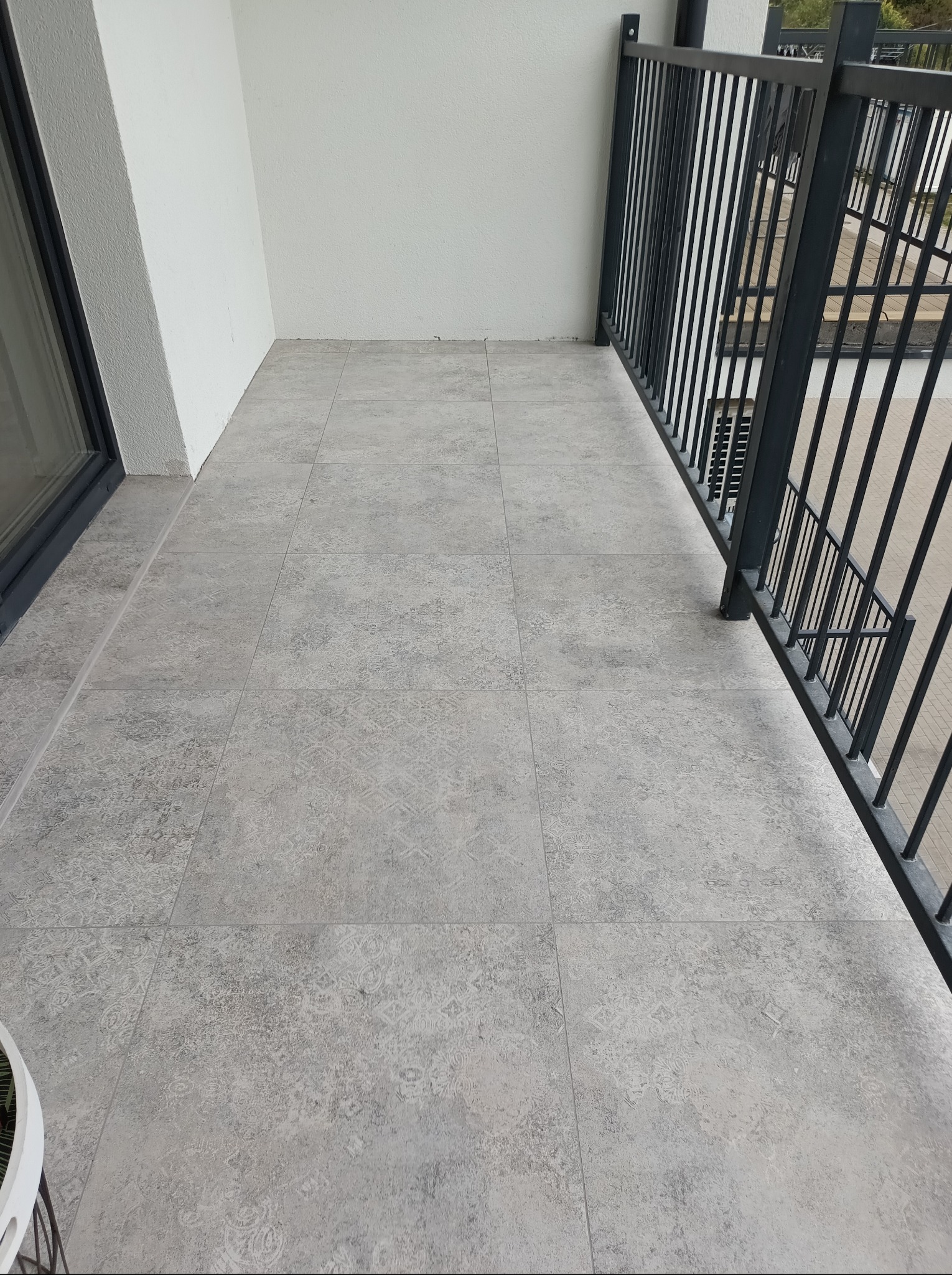 Wykończony balkon z szarymi płytkami o wzorzystej fakturze. Czarne, metalowe barierki. Świeżo pomalowane, białe ściany. Widok z góry, brak elementów dekoracyjnych.