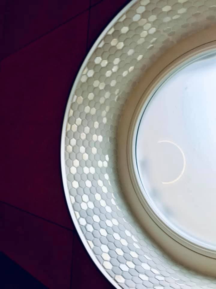 Okrągłe lustro z dekoracyjną ramą z heksagonalnych płytek w odcieniach szarości i bieli, na tle bordowej ściany. Nietypowe ujęcie z bliska, oryginalny design.