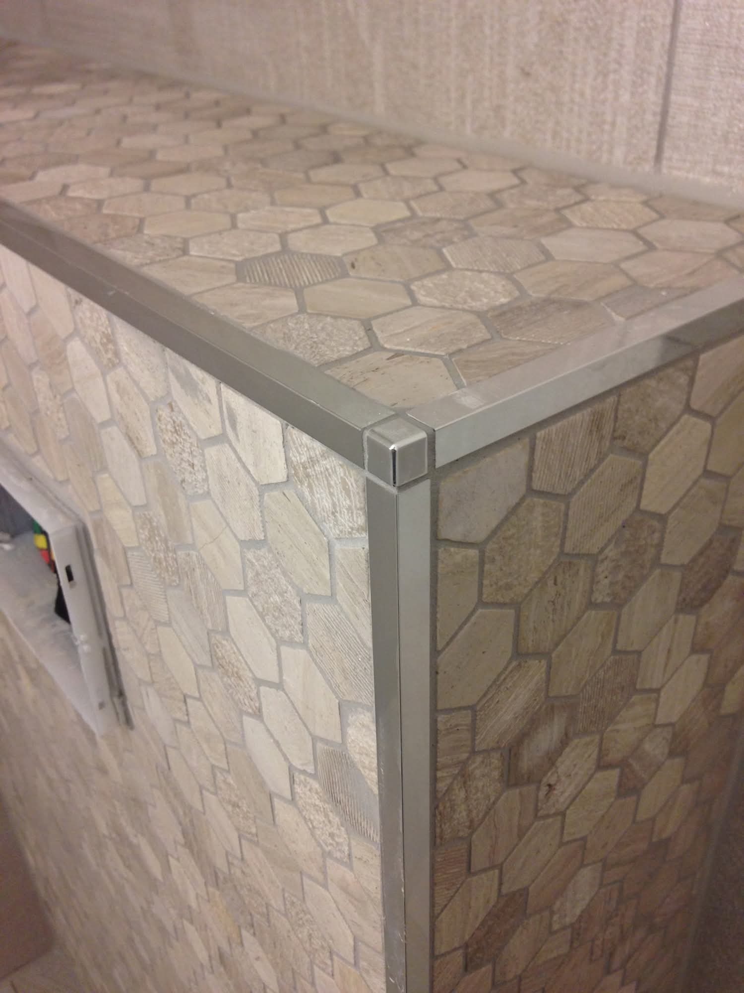 Wykończona obudowa z heksagonalnych płytek w odcieniach beżu, zabezpieczona srebrną listwą narożną, ukazana w zbliżeniu.