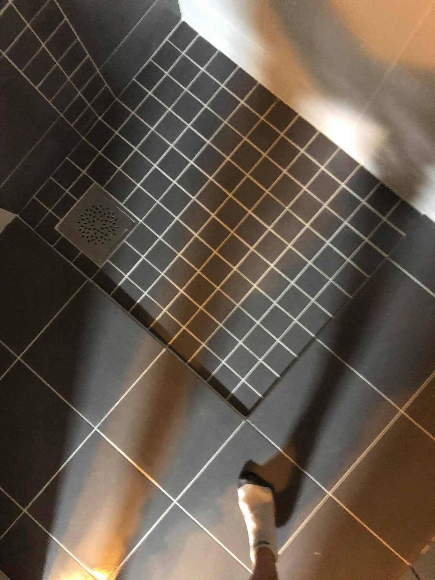 Ciemnoszara, kwadratowa mozaika podłogowa w kabinie prysznicowej z odpływem. Na podłodze widoczna stopa w skarpetce. Ujęcie z góry.