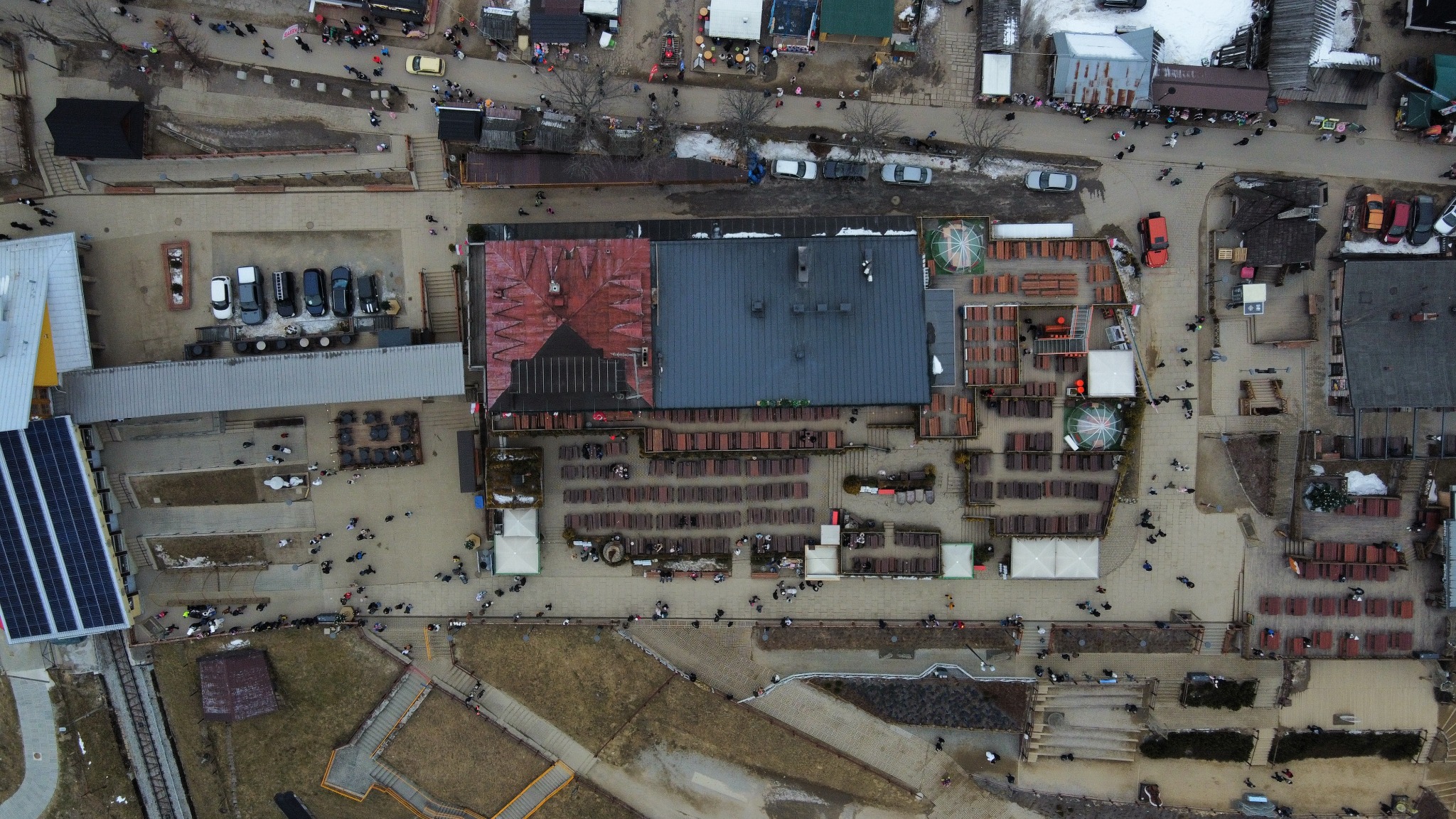 Widok z lotu ptaka na kompleks budynków z restauracją i miejscami siedzącymi na zewnątrz, otoczony ulicami i chodnikami z przechodzącymi ludźmi. Obraz z drona.