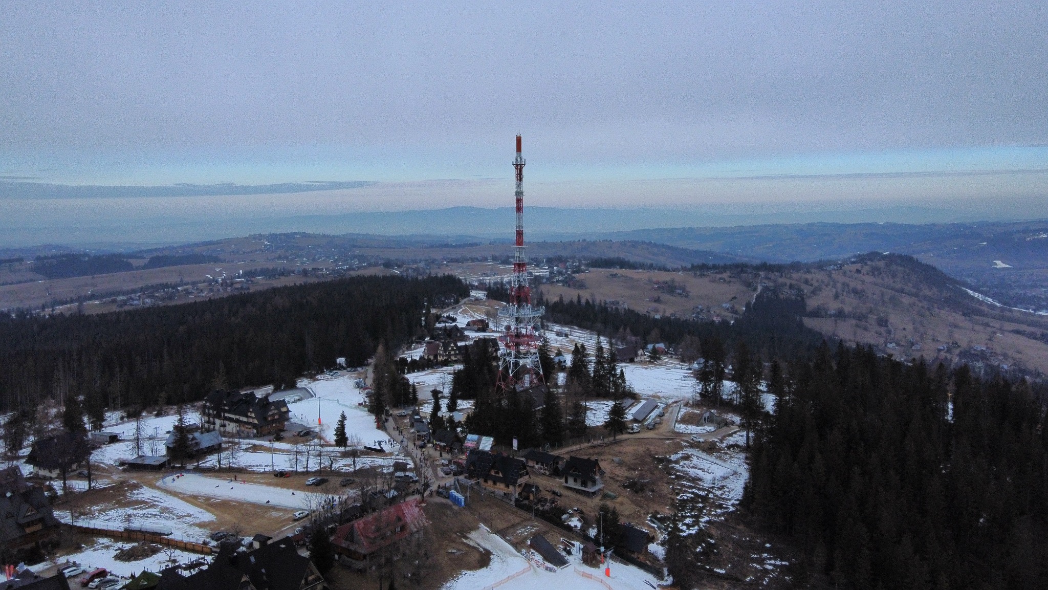 Widok z lotu ptaka na wieżę nadawczą na szczycie góry w zimowej scenerii. W tle widać zalesione wzgórza i zabudowania.