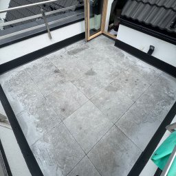 Roof-Bud - Płaski dach z szarymi płytkami, otwartym oknem dachowym i fragmentem czarnego dachu z dachówkami. Widoczne ślady po wodzie na płytkach.