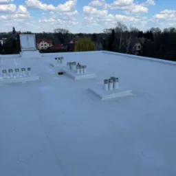 Biały, płaski dach pokryty membraną, z kominami wentylacyjnymi i wyłazem dachowym na tle zabudowy miejskiej i błękitnego nieba z chmurami.