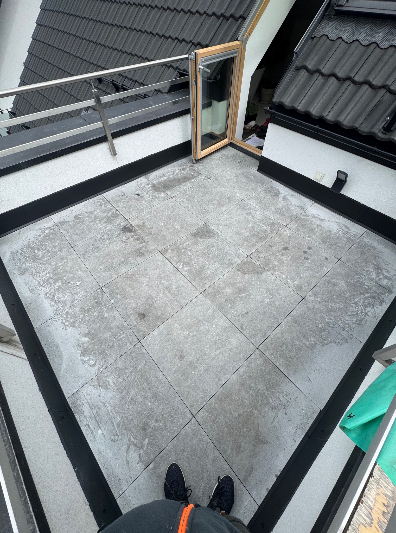 Płaski dach z szarymi płytkami, otwartym oknem dachowym i fragmentem czarnego dachu z dachówkami. Widoczne ślady po wodzie na płytkach.