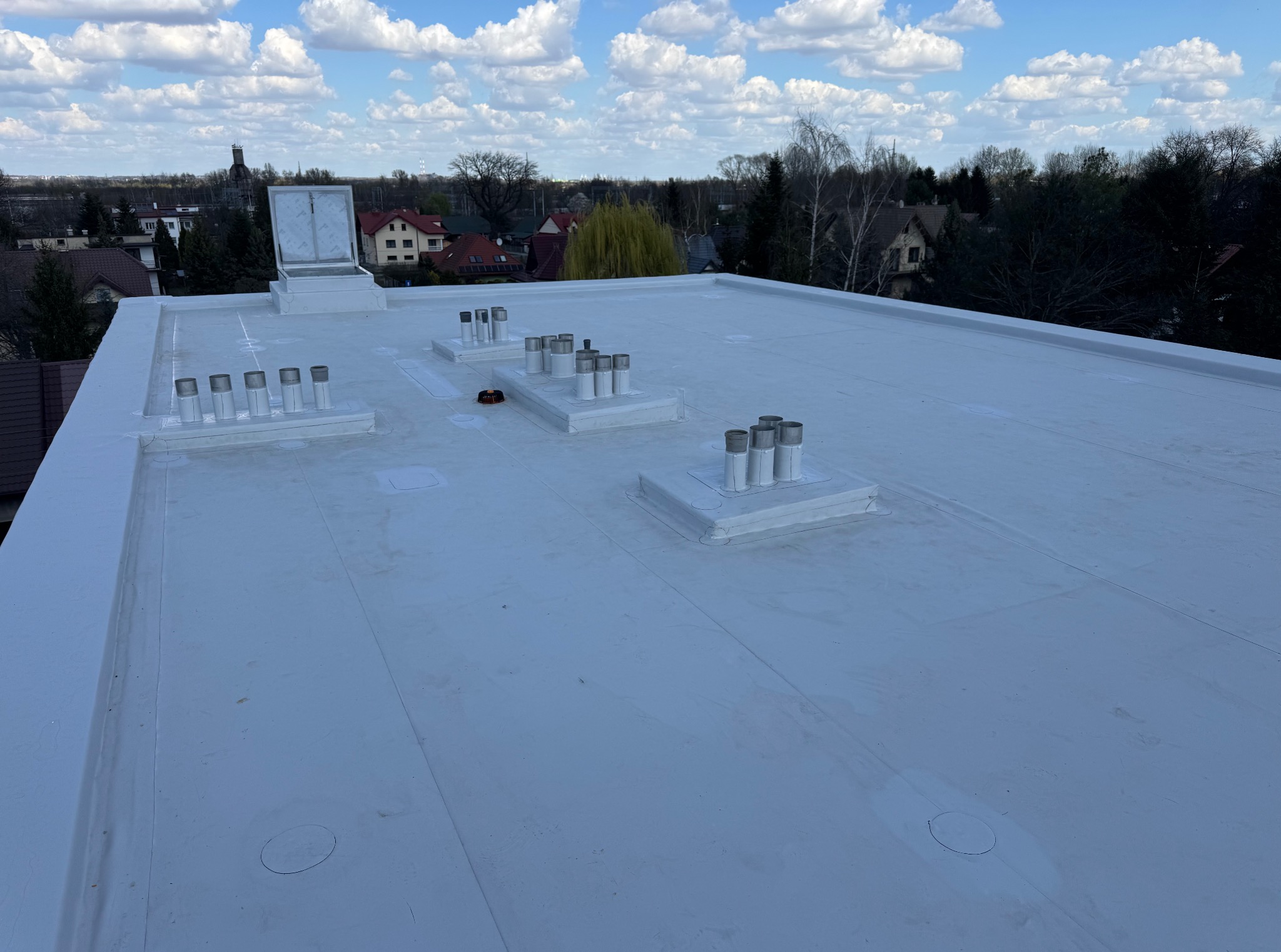 Biały, płaski dach pokryty membraną, z kominami wentylacyjnymi i wyłazem dachowym na tle zabudowy miejskiej i błękitnego nieba z chmurami.