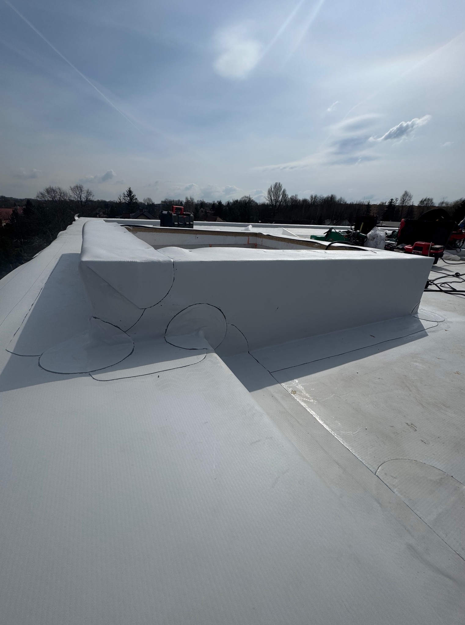 Biała, płaska powierzchnia dachu z obróbkami blacharskimi i narzędziami w tle, widziana z perspektywy dachu w słoneczny dzień.