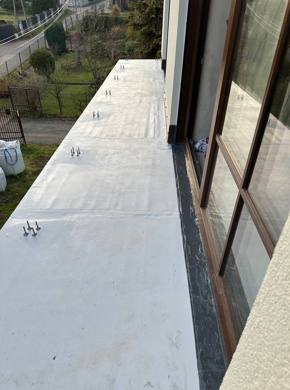 Biała, płaska powierzchnia dachu pokryta papą, z widocznymi mocowaniami i oknem w tle. Widok z góry.