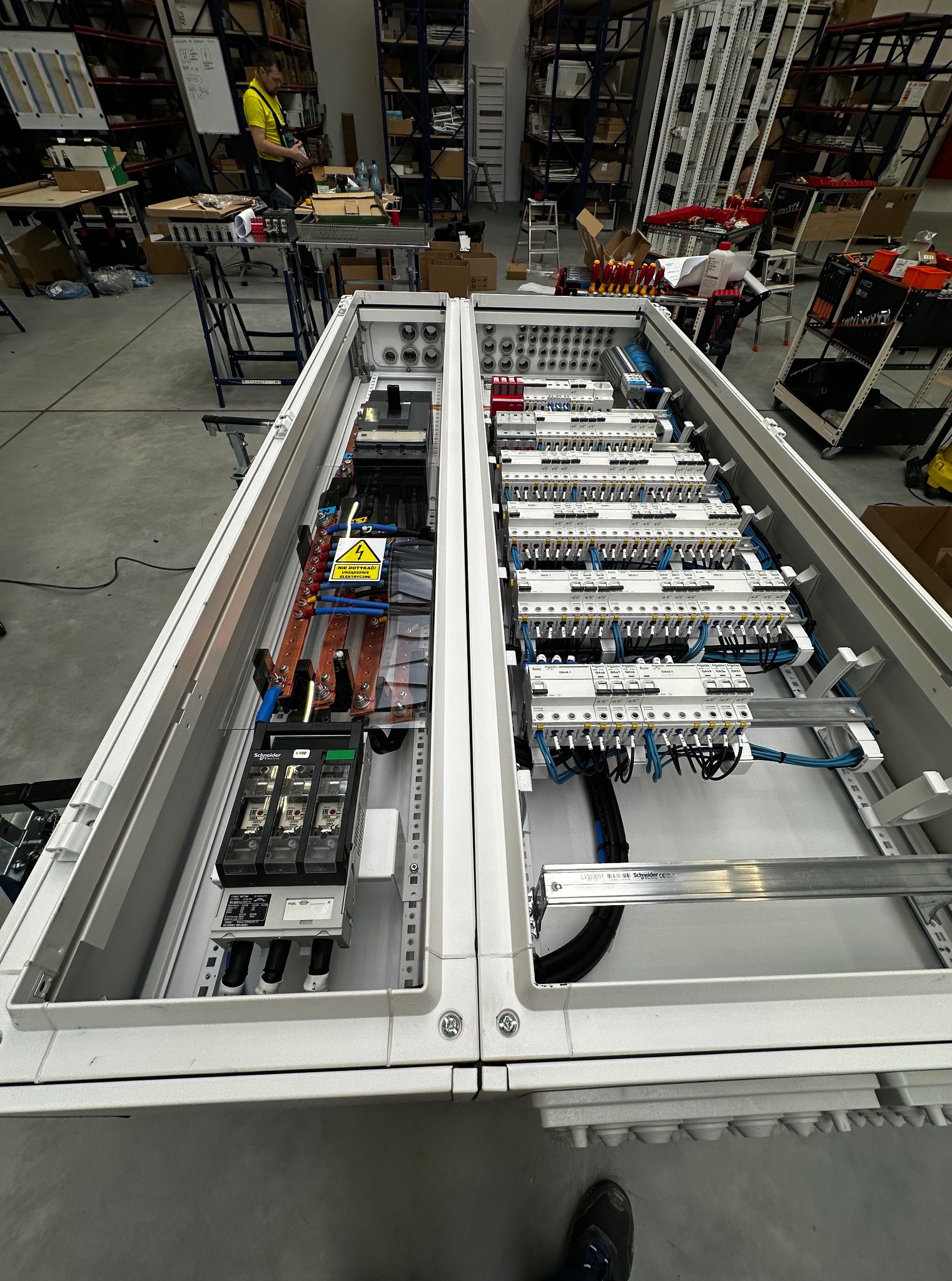 Otwarta szafa elektryczna z widocznymi bezpiecznikami, przewodami i złączkami. Szczegółowe okablowanie wewnątrz, w tle magazyn z regałami.