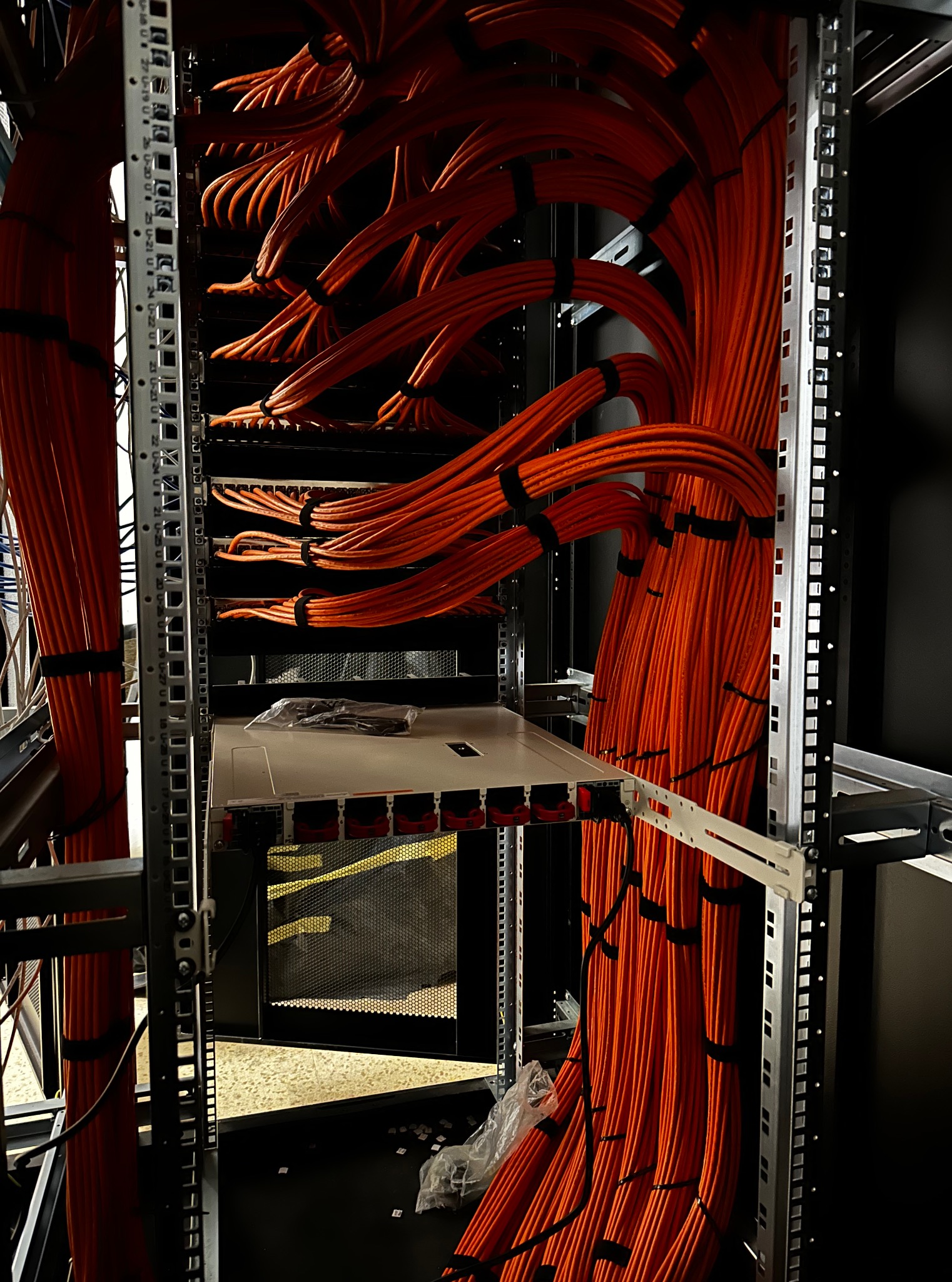 Pomarańczowe kable sieciowe ułożone w szafie rack, spięte czarnymi opaskami. Widoczny fragment serwera i metalowa konstrukcja szafy. Oświetlenie górne.