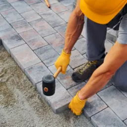 Pracownik w kasku i rękawicach układa kostkę brukową z użyciem gumowego młotka. Częściowo ułożona nawierzchnia z kostki o szaro-ceglastej barwie.