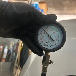 Marcin Podraza - Manometr Geko w czarnej rękawicy, wskazujący ciśnienie w samochodzie. W tle widoczne akcesoria warsztatowe. Pomiar ciśnienia oleju w silniku.