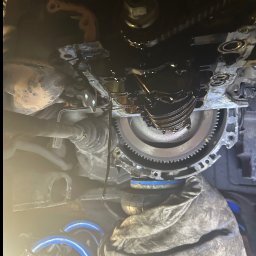 Marcin Podraza - Silnik samochodowy w trakcie naprawy, widoczne elementy mechaniczne i wyciekający olej. Ujęcie z dołu, praca w warsztacie.