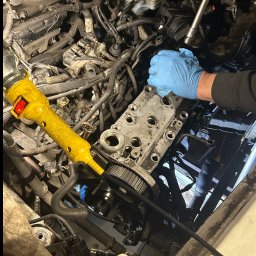 Marcin Podraza - Silnik samochodowy w trakcie naprawy z widoczną głowicą cylindrów, ręką w niebieskiej rękawicy i narzędziem. Żółta lampa warsztatowa oświetla obszar roboczy.