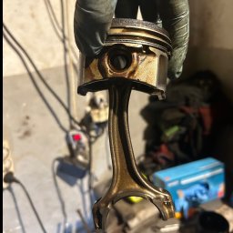 Marcin Podraza - Tłok silnika spalinowego, trzymany w dłoni w rękawicy roboczej, widoczne ślady zużycia i nagaru, w tle warsztat samochodowy.