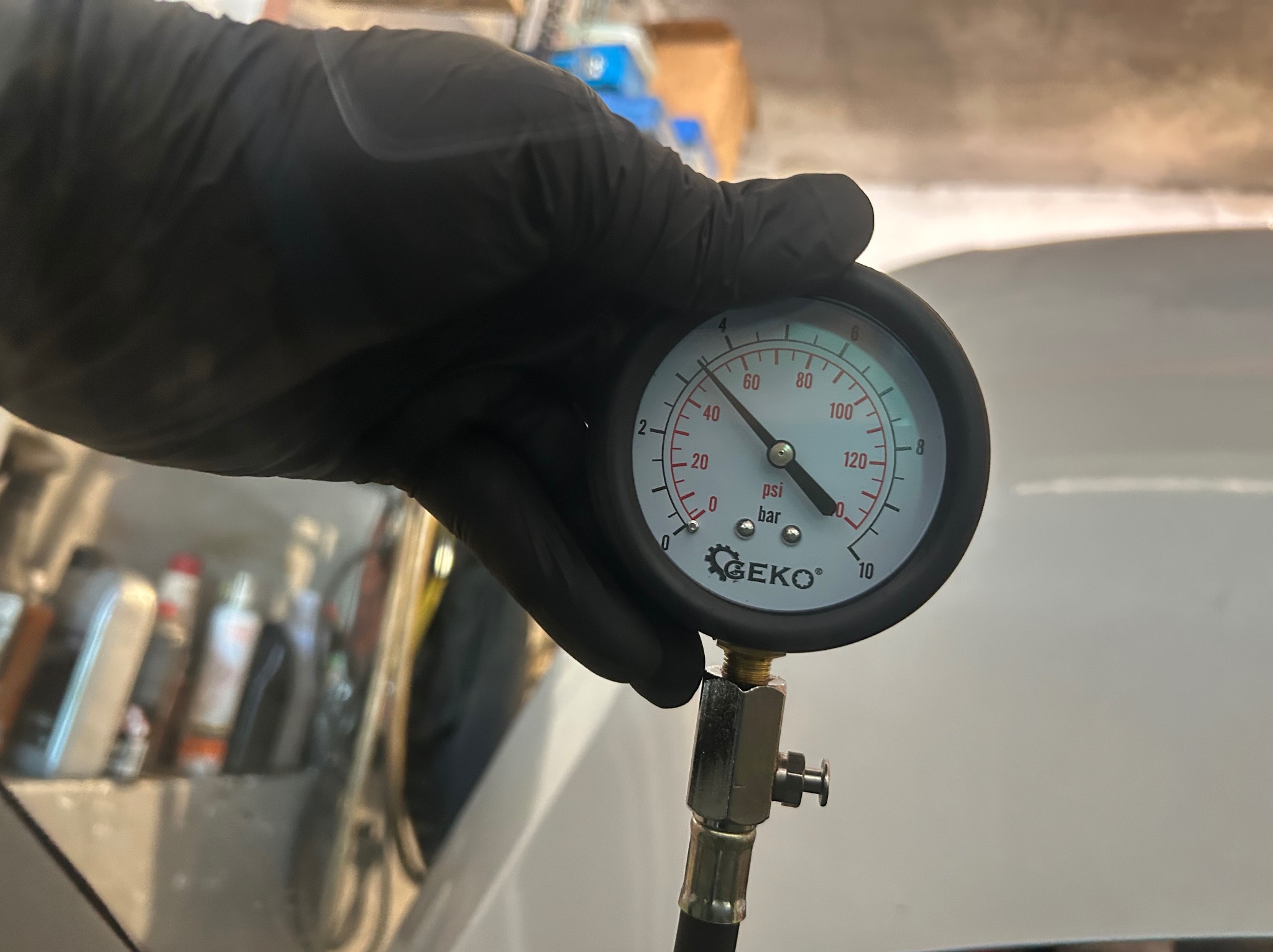 Manometr Geko w czarnej rękawicy, wskazujący ciśnienie w samochodzie. W tle widoczne akcesoria warsztatowe. Pomiar ciśnienia oleju w silniku.