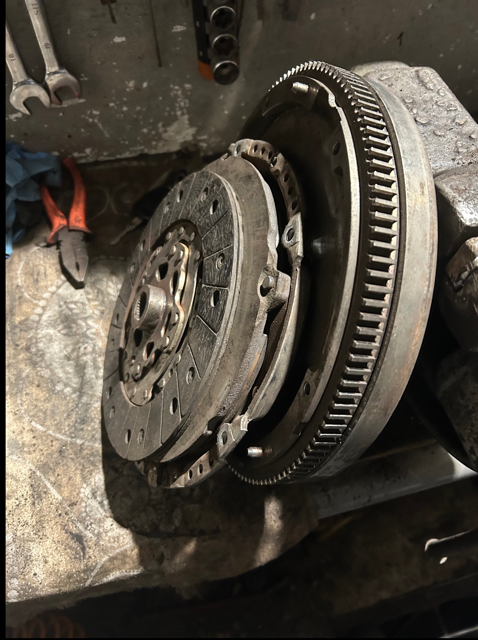 Sprzęgło samochodowe z kołem zamachowym na brudnym stole warsztatowym, obok narzędzia: klucze i kombinerki. Widoczne ślady użytkowania.