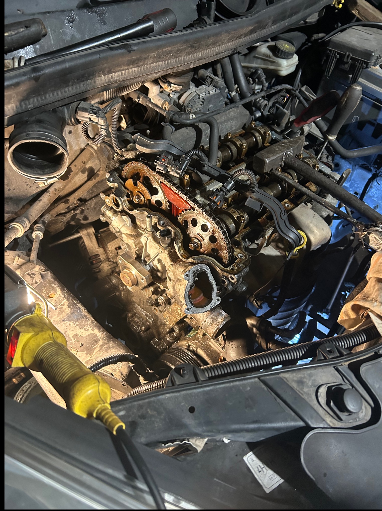 Otwarty silnik samochodowy z widocznym łańcuchem rozrządu, oświetlony żółtą lampą warsztatową. Detal mechaniczny w trakcie naprawy lub serwisu.