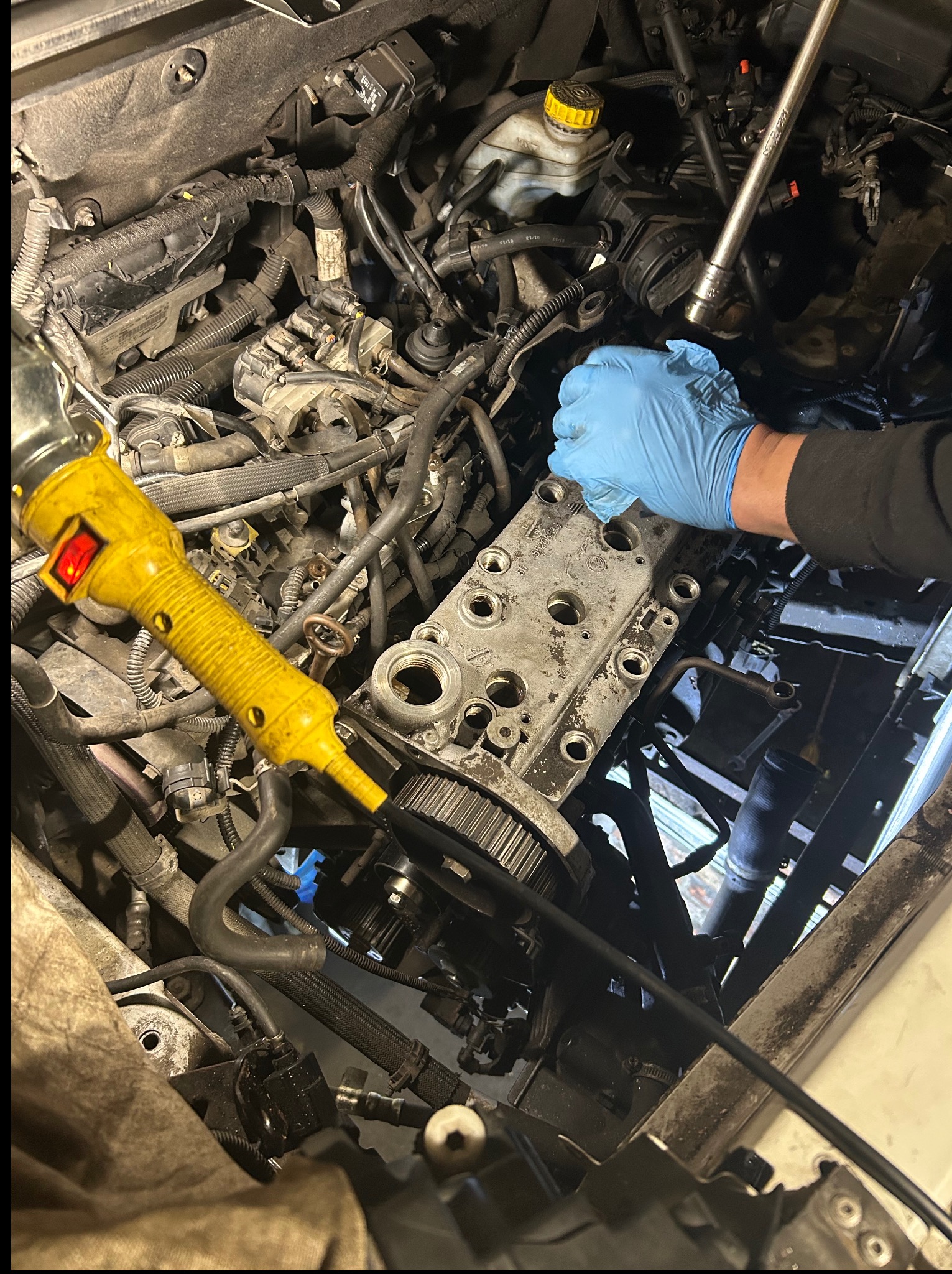 Silnik samochodowy w trakcie naprawy z widoczną głowicą cylindrów, ręką w niebieskiej rękawicy i narzędziem. Żółta lampa warsztatowa oświetla obszar roboczy.