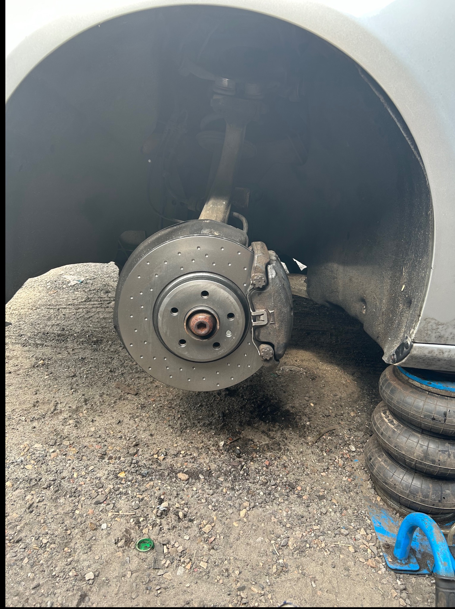 Nowa tarcza hamulcowa z zaciskiem w srebrnym aucie, widoczna w nadkolu. Obok leżą opony i podnośnik pneumatyczny. Tarcza posiada nawiercenia dla lepszego chłodzenia.