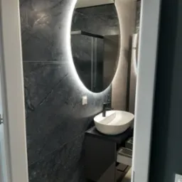 Nowoczesna łazienka z owalnym lustrem LED, umywalką nablatową i ścianą wyłożoną płytkami imitującymi ciemny marmur. Minimalistyczny design i eleganckie wykończenie.