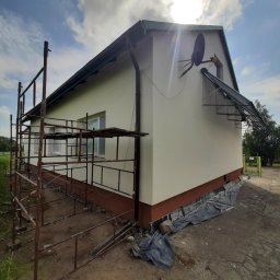 Łukasz Piasecki - Dom po malowaniu elewacji, widoczne rusztowania i nowa, jasna kolorystyka ścian. Ciemna podmurówka i antena satelitarna na świeżo pomalowanej fasadzie.