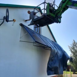 Łukasz Piasecki - Pracownik na podnośniku maluje elewację domu. Widoczny fragment elewacji z osłoniętym kolektorem słonecznym. Słoneczny dzień.